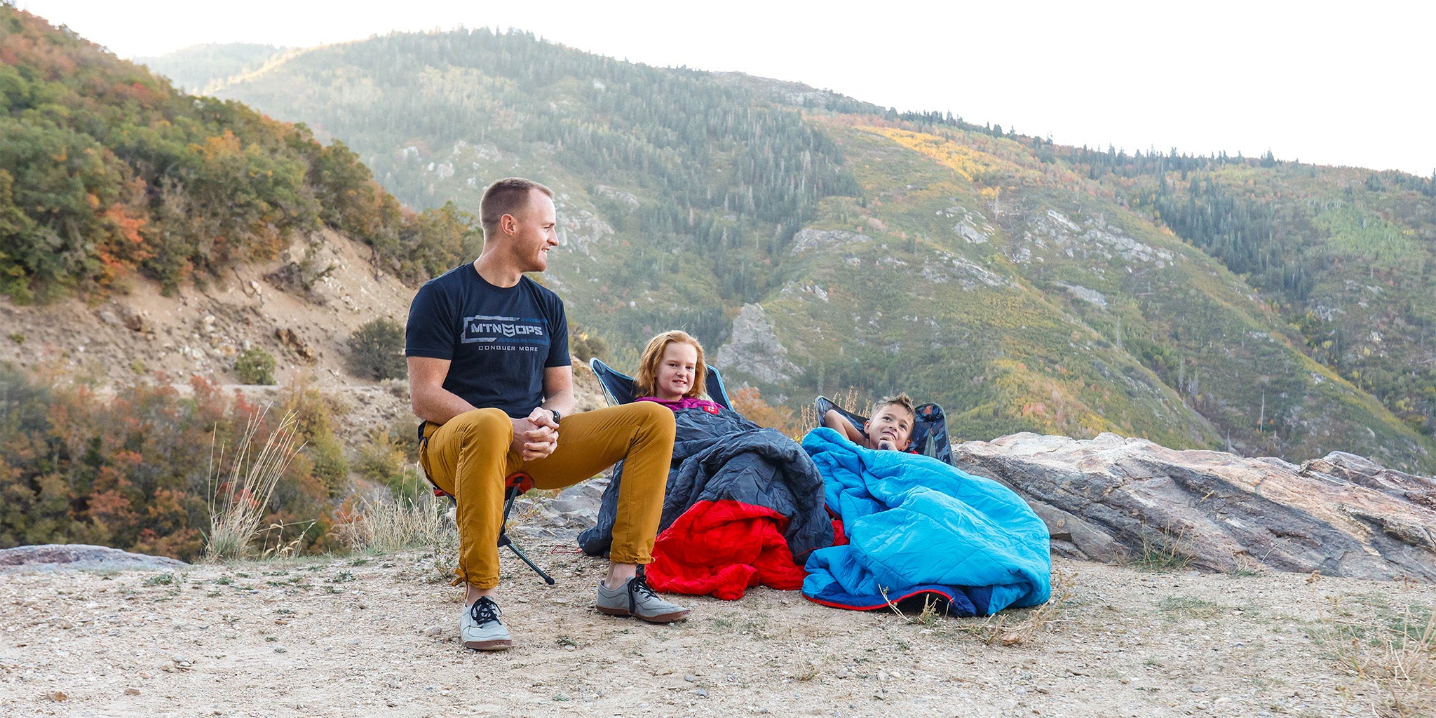 Man sits on compass 360 stool while children sit in chairs with thermaquilts mountain background fall foliage