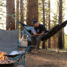 Man lounging in evolution hammock eating mountain house meals by a firepit with his dog looking longingly for some food